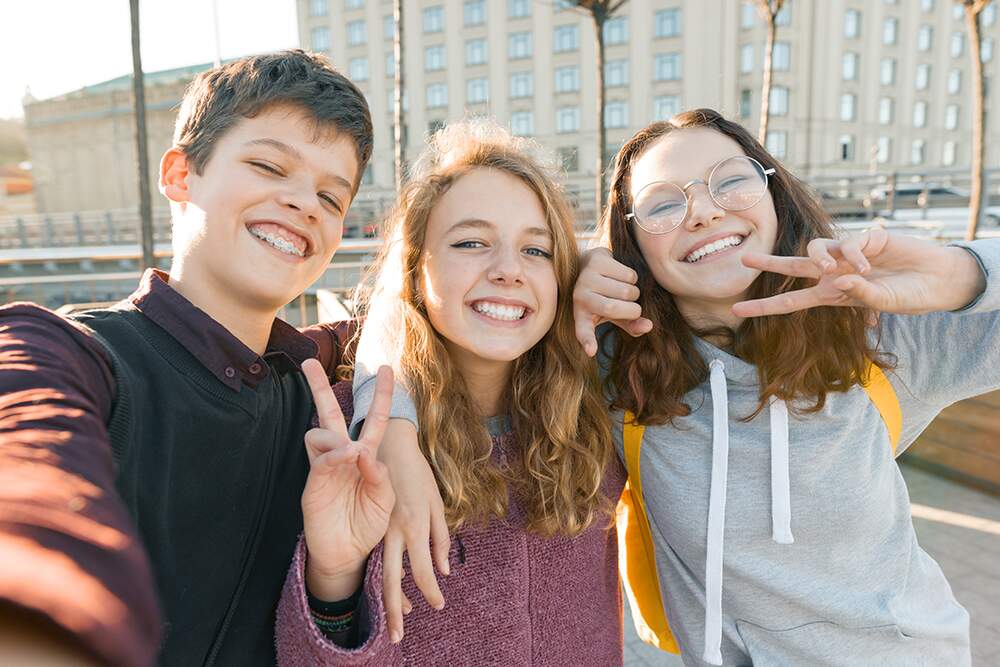 Portrait of three teen friends boy and two girls smiling and taking a selfie outdoors. City background, golden hour.